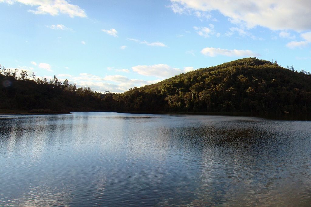 Lago de águas calmas refletindo o céu azul, cercado por morros cobertos de mata