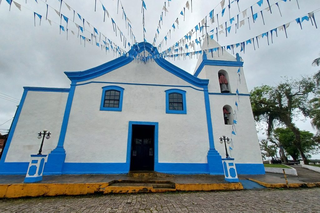 Fachada branca com detalhes azuis da Igreja Matriz São João Batista de Cananéia, enfeitada com bandeirinhas