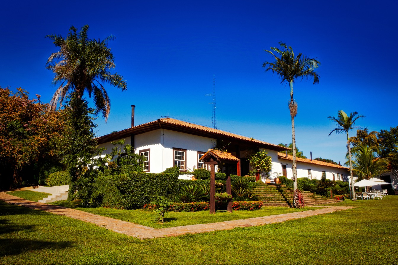 Casa-sede branca da Fazenda Capoava, com janelas de madeira e telhado de telhas, entre palmeiras e jardim gramado sob céu azul