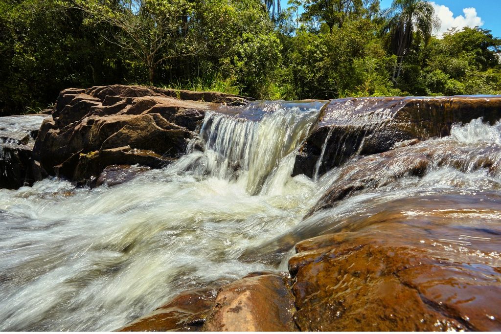Água correndo sobre lajes de pedra avermelhada na Cachoeira do Cubatão, em Cássia dos Coqueiros, com mata ao fundo