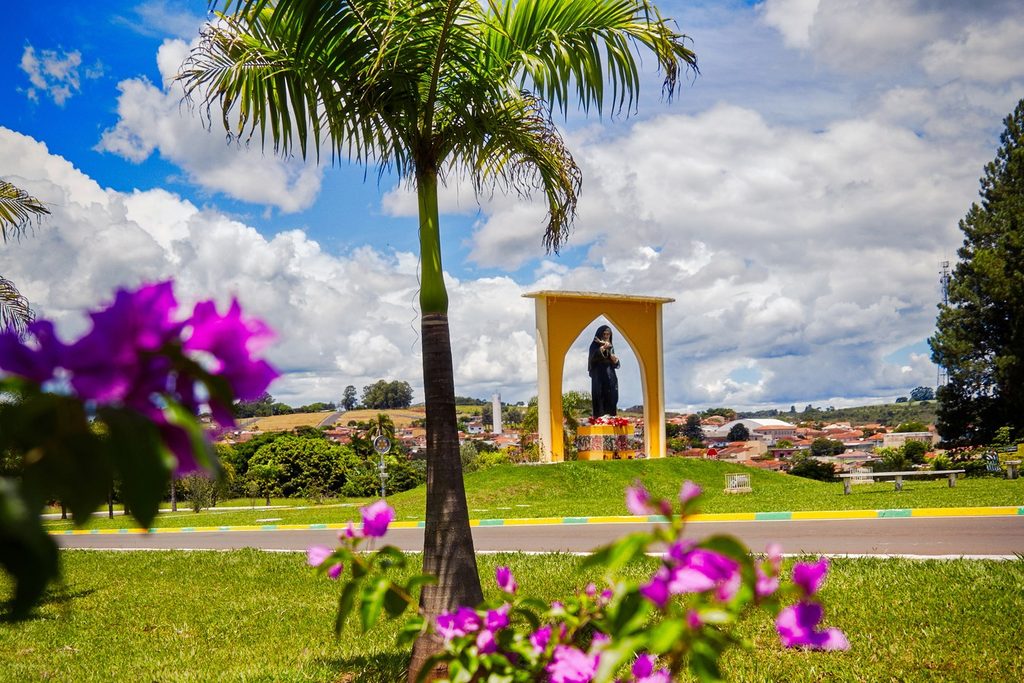Portal amarelo com a imagem de Santa Rita de Cássia na entrada da cidade, com palmeira e flores roxas em primeiro plano