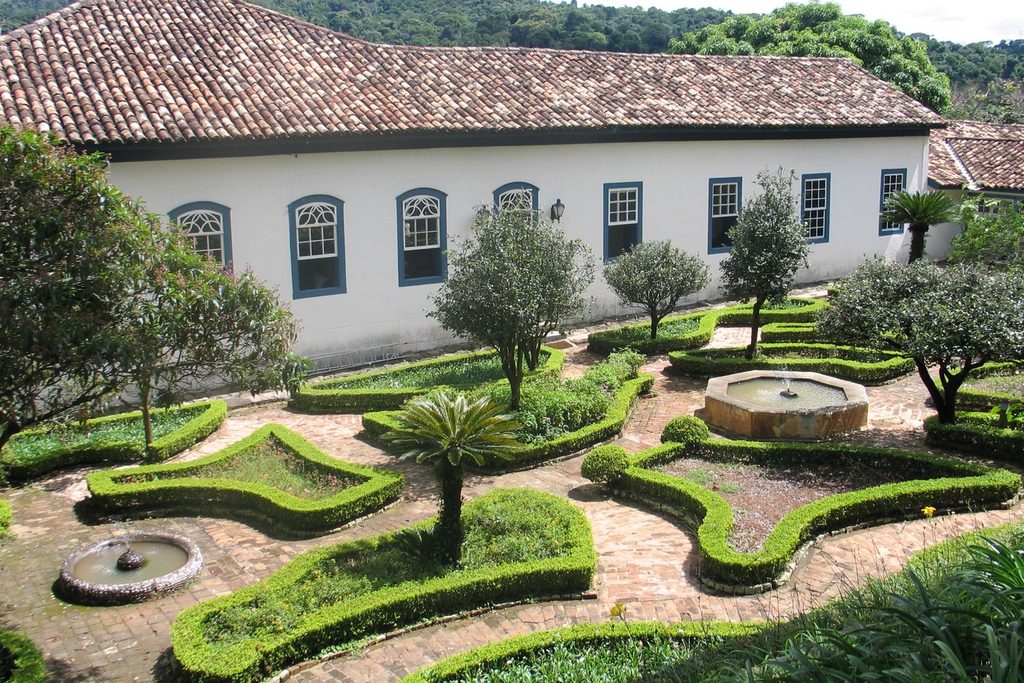 Jardim com canteiros de buxinho, chafariz e oliveiras diante da ala comprida do casarão da Fazenda Dona Carolina