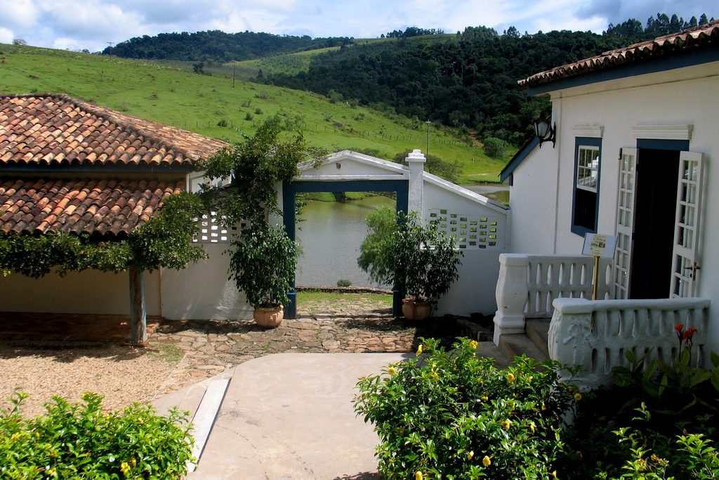 Pátio de pedras da Fazenda Dona Carolina com construções coloniais brancas, vasos de plantas e o lago ao fundo entre morros verdes