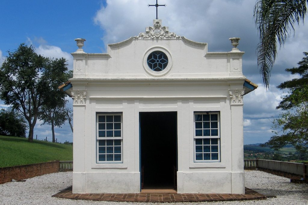 Capela branca de fachada ornamentada, com janela redonda, cruz no alto e porta aberta, num terraço de cascalho