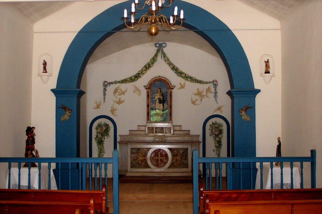 Interior de capela antiga com arco azul, altar decorado, imagens de santos e lustre de velas