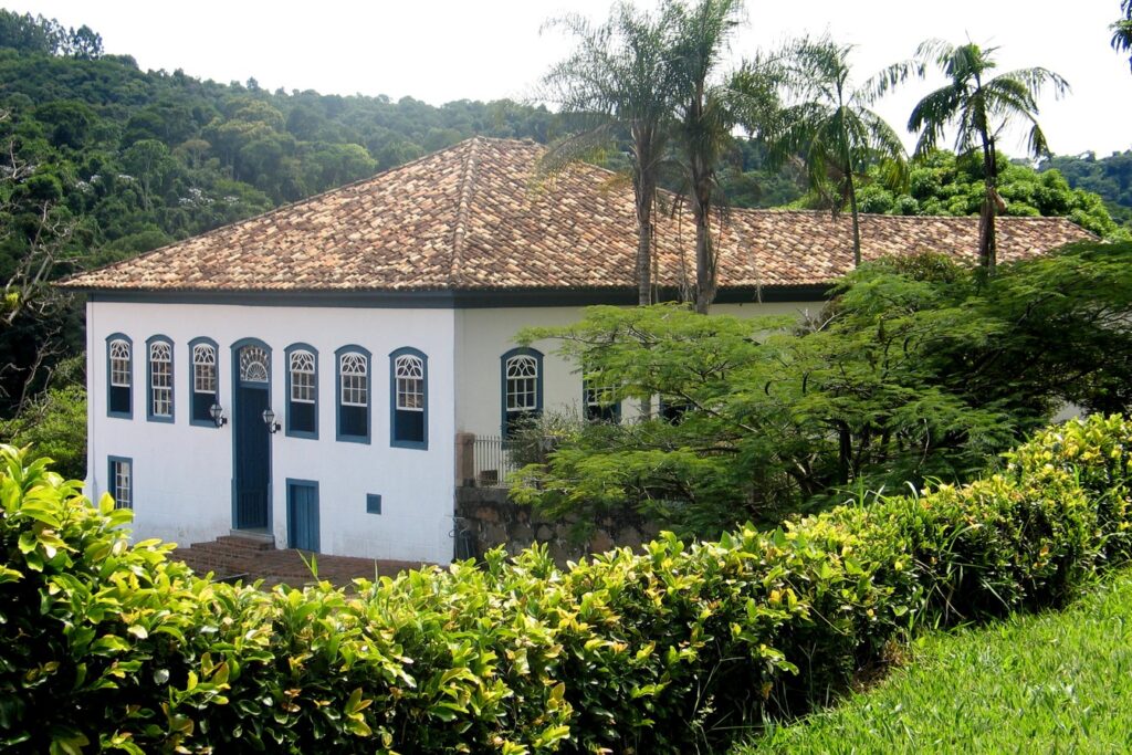 Casarão colonial branco de dois andares da Fazenda Dona Carolina, com janelas azuis e telhado de barro, entre palmeiras e mata