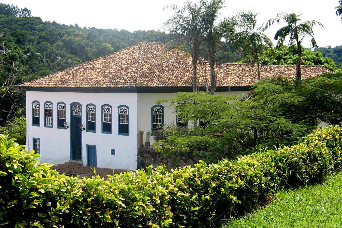 Casarão colonial branco de dois andares da Fazenda Dona Carolina, com janelas azuis e telhado de barro, entre palmeiras e mata