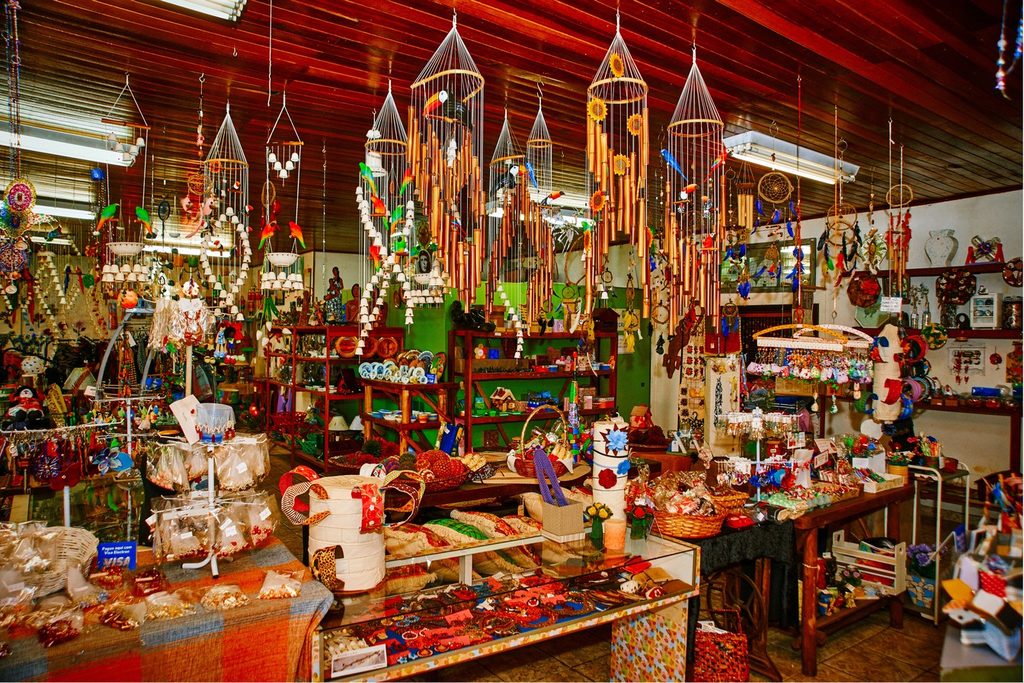 Loja cheia de artesanato colorido, com mensageiros do vento pendurados no teto e peças sobre as mesas, na Casa do Artesão