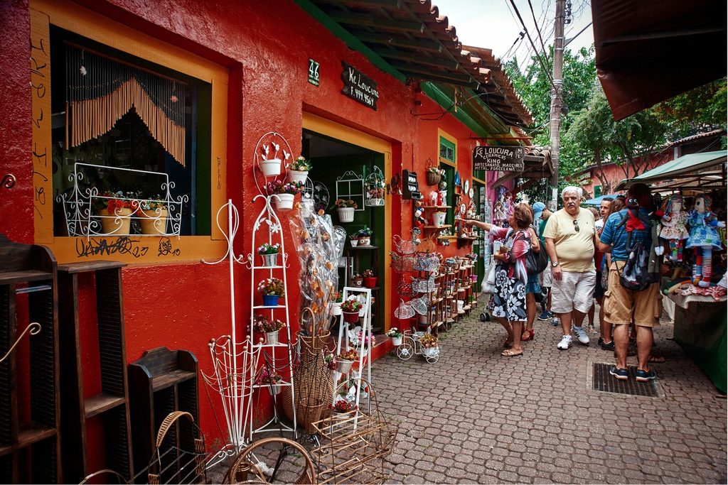 Rua de pedra com lojas coloridas e peças de artesanato expostas na calçada, cheia de visitantes, durante a feira de Embu das Artes