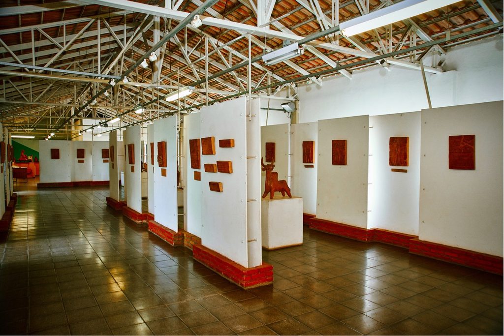 Galeria com paredes brancas e painéis expondo peças de cerâmica de Tadakiyo Sakai, sob telhado de estrutura metálica
