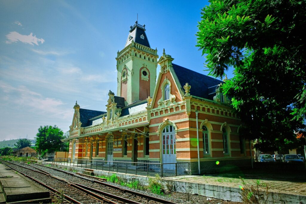 Estação ferroviária de Guaratinguetá, de tijolos vermelhos e faixas claras, com torre central de relógio e telhado de ardósia, ao lado dos trilhos