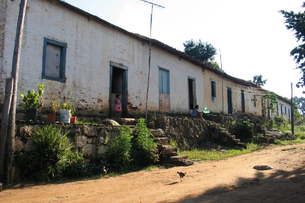 Fileira de casas antigas de colonos com portas e janelas azuis, à beira de uma rua de terra