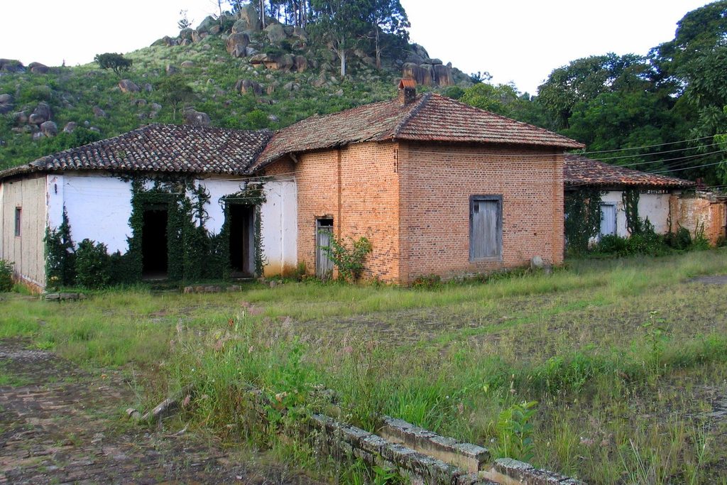 Conjunto de construções antigas da fazenda, uma caiada de branco e outra de tijolo aparente, com morro de pedras ao fundo