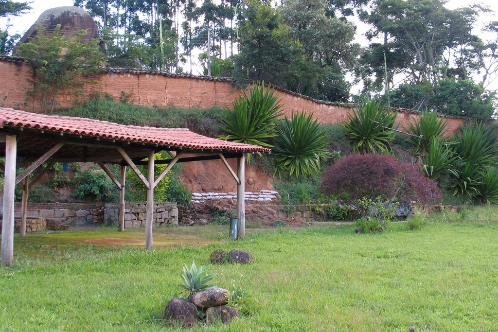 Quiosque de telhado vermelho num gramado com palmeiras e um muro alto de terra ao fundo