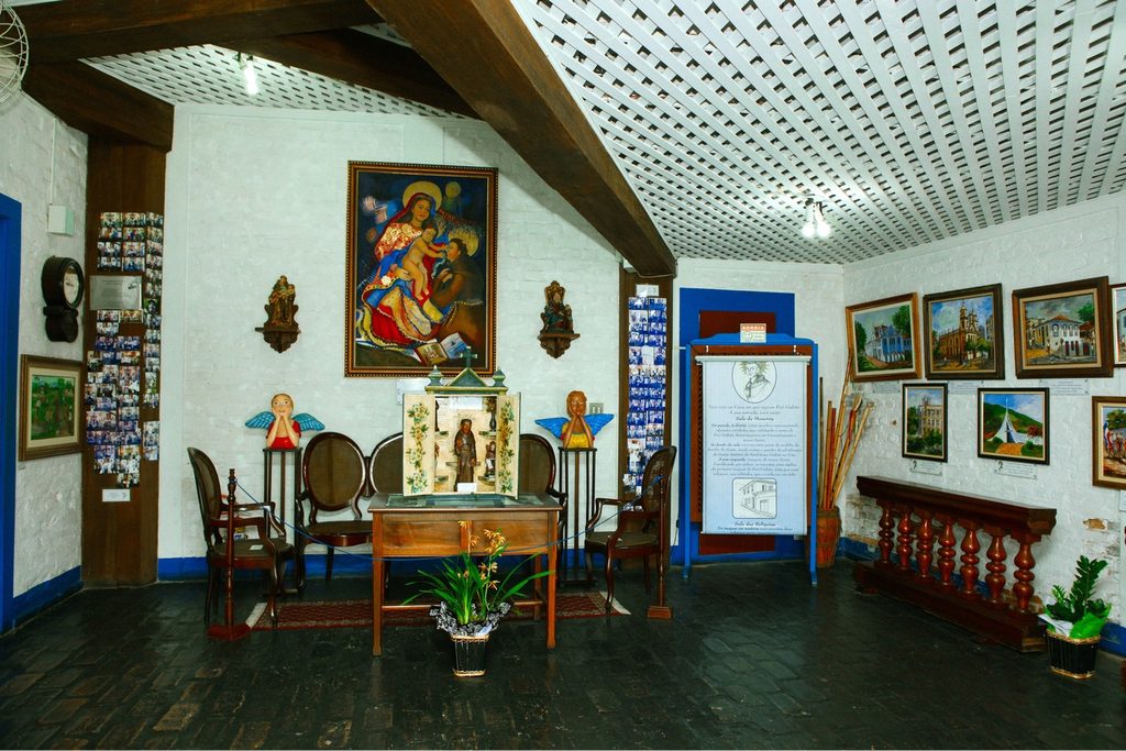 Sala da Casa de Frei Galvão com quadros religiosos, imagens de santos, mesa de madeira, cadeiras antigas e painel com a história do santo