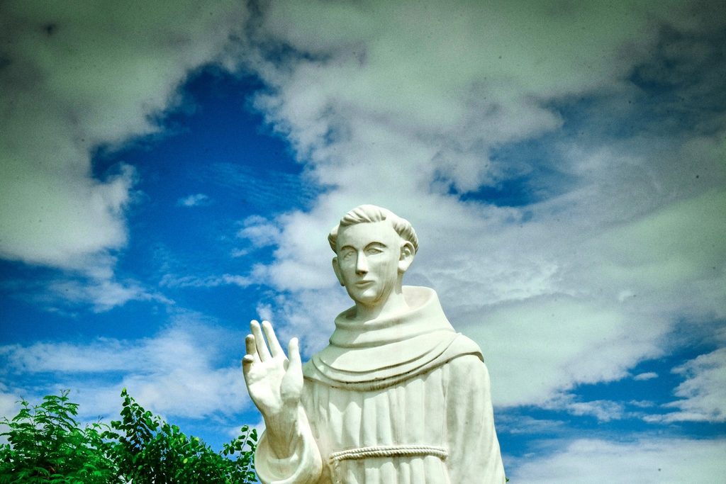 Estátua branca de Frei Galvão com hábito franciscano e a mão erguida, contra o céu azul com nuvens