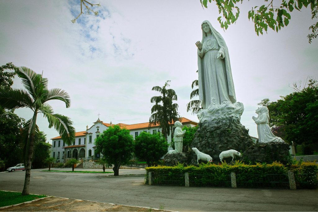 Estátua gigante de Nossa Senhora de Fátima com os pastorinhos e ovelhas diante do prédio do seminário franciscano, entre palmeiras