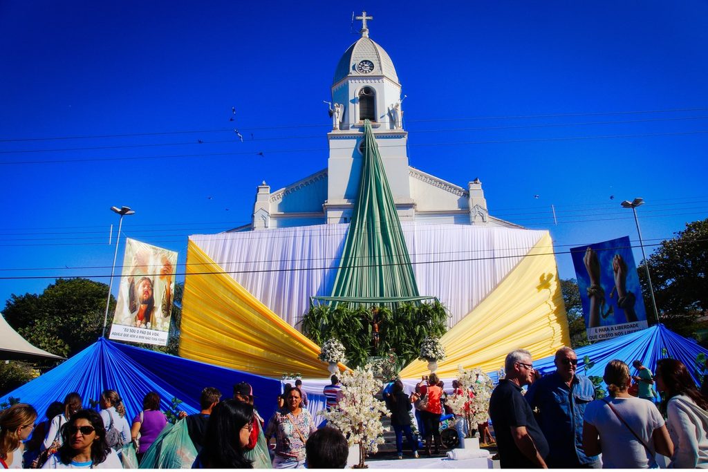 Fiéis em frente à igreja enfeitada com grandes tecidos azuis, amarelos e verdes na festa de Corpus Christi