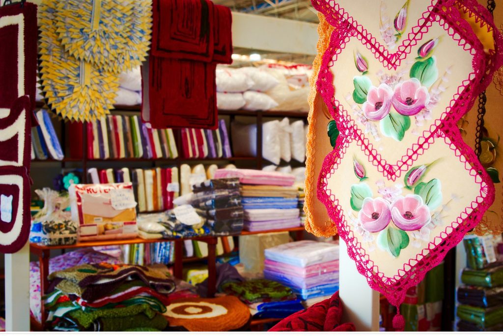 Panos de prato bordados com rosas cor-de-rosa e prateleiras cheias de toalhas e enxovais coloridos numa banca da feira de Ibitinga