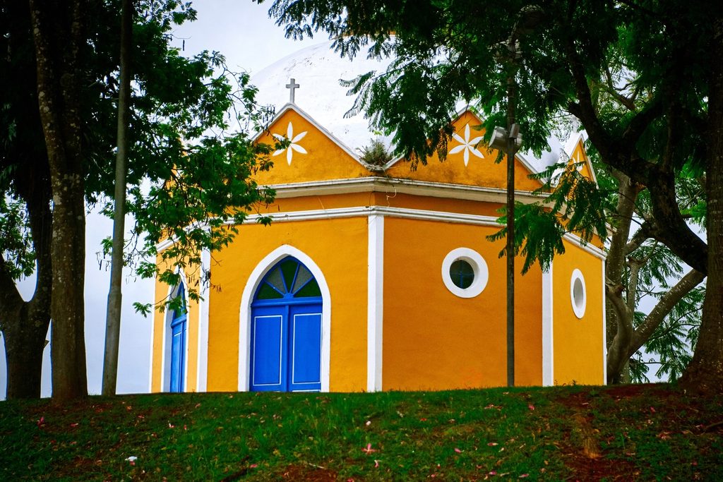 Capela do Bom Jesus da Prisão, em Ibiúna, amarela com porta azul em arco e cruz no alto, entre árvores