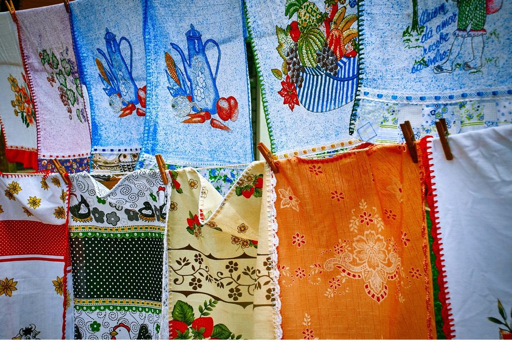 Panos de prato bordados e pintados com bules, frutas e flores pendurados num varal da feira de artesanato