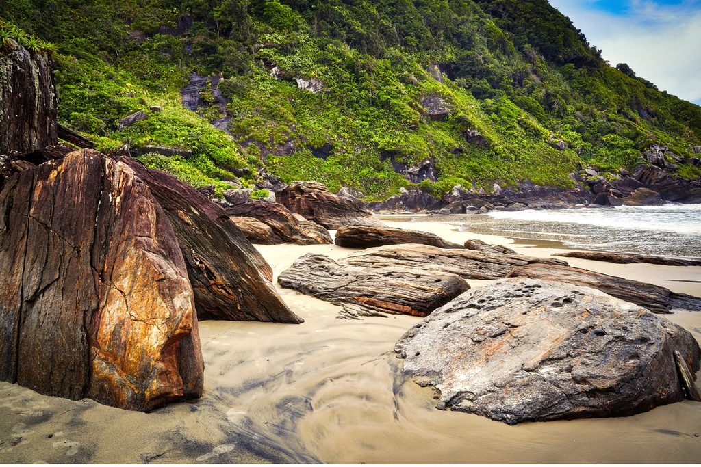 Grandes pedras escuras na areia do Costão da Juréia, com mar e encosta coberta de Mata Atlântica