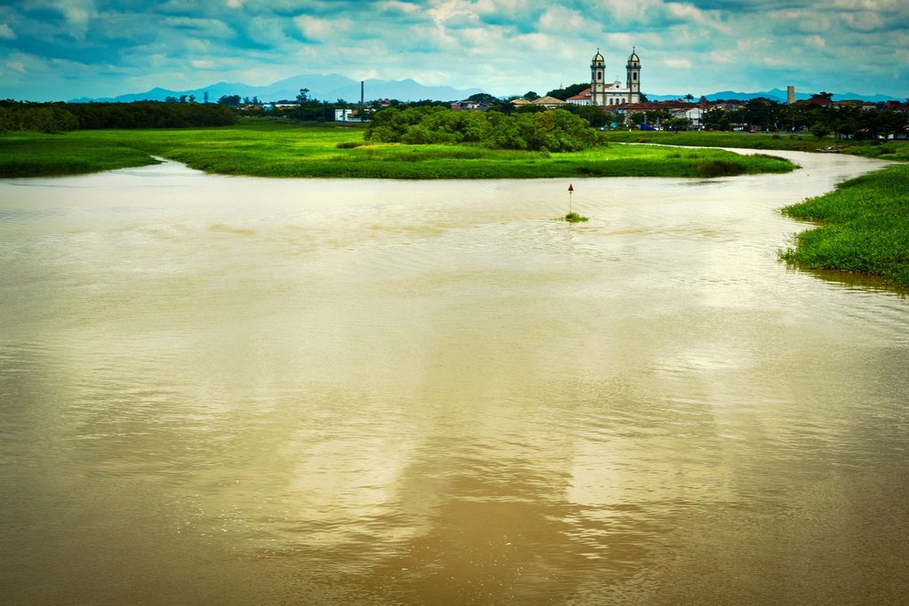 Águas largas do estuário cercadas de vegetação, com as torres da basílica ao fundo