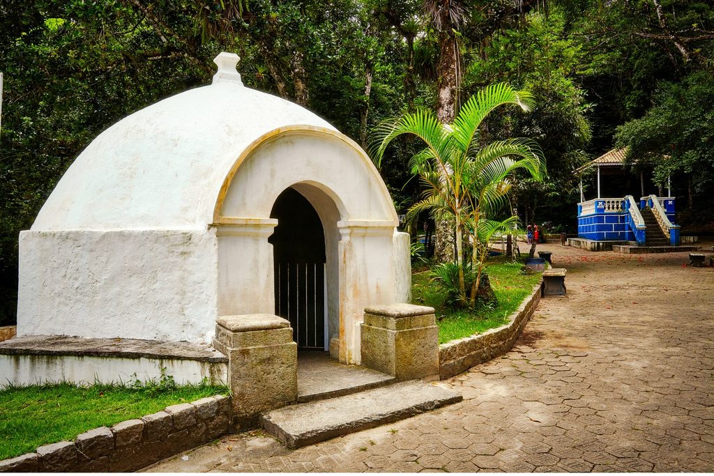 Pequena construção branca em forma de cúpula sobre a Fonte do Senhor, com caminho de pedras e mata ao redor