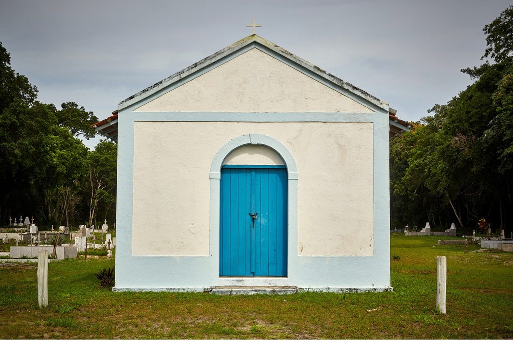 Capela branca simples com porta azul em arco, cercada de árvores, na Ilha Comprida
