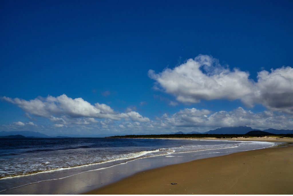Praia longa e quase deserta, com ondas suaves e céu azul com nuvens, na Ponta da Praia da Ilha Comprida
