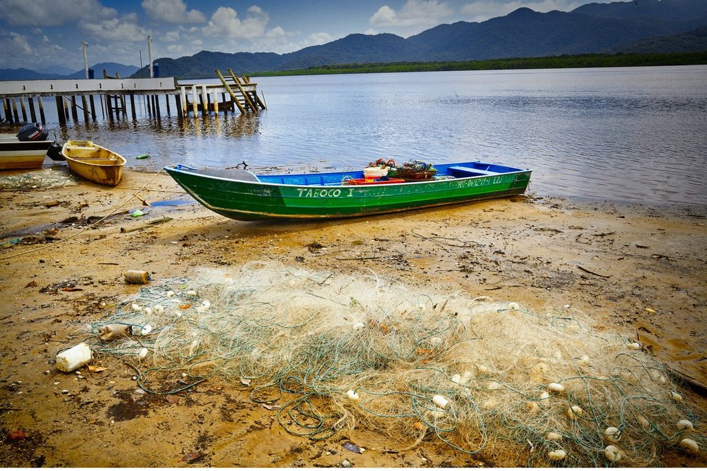 Barco de pesca verde na areia ao lado de uma rede, com píer de madeira e serras ao fundo, na Vila de Pedrinhas