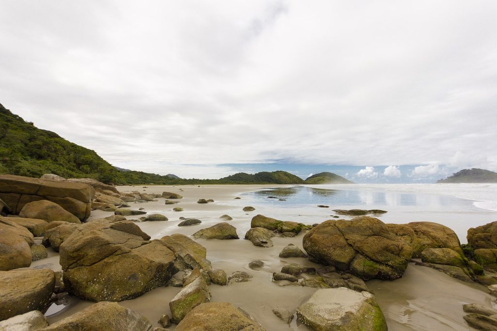 Pedras arredondadas na areia molhada da Praia do Cambriú, com morros cobertos de mata ao fundo