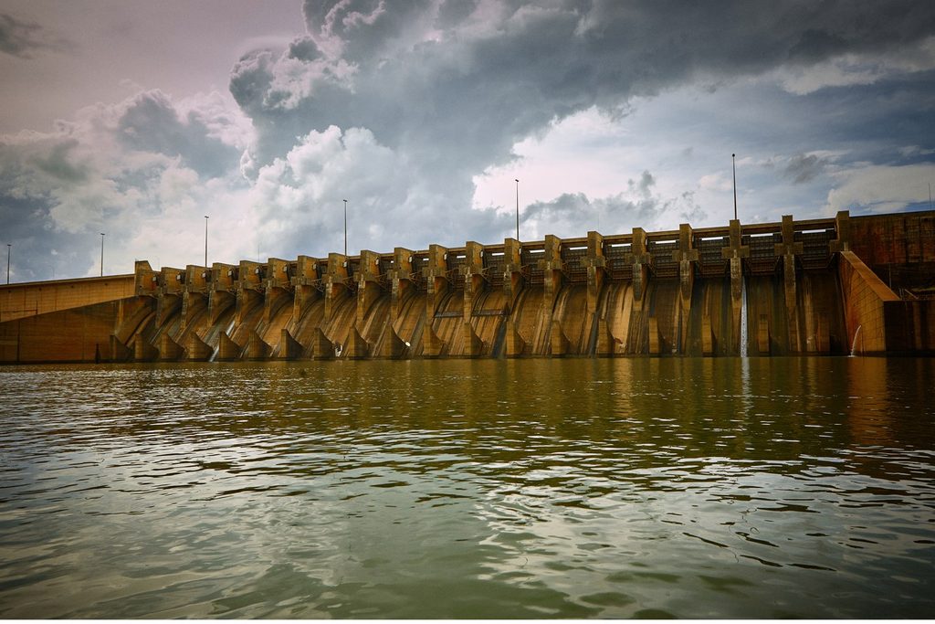 Barragem da usina hidrelétrica vista da água, sob céu carregado de nuvens