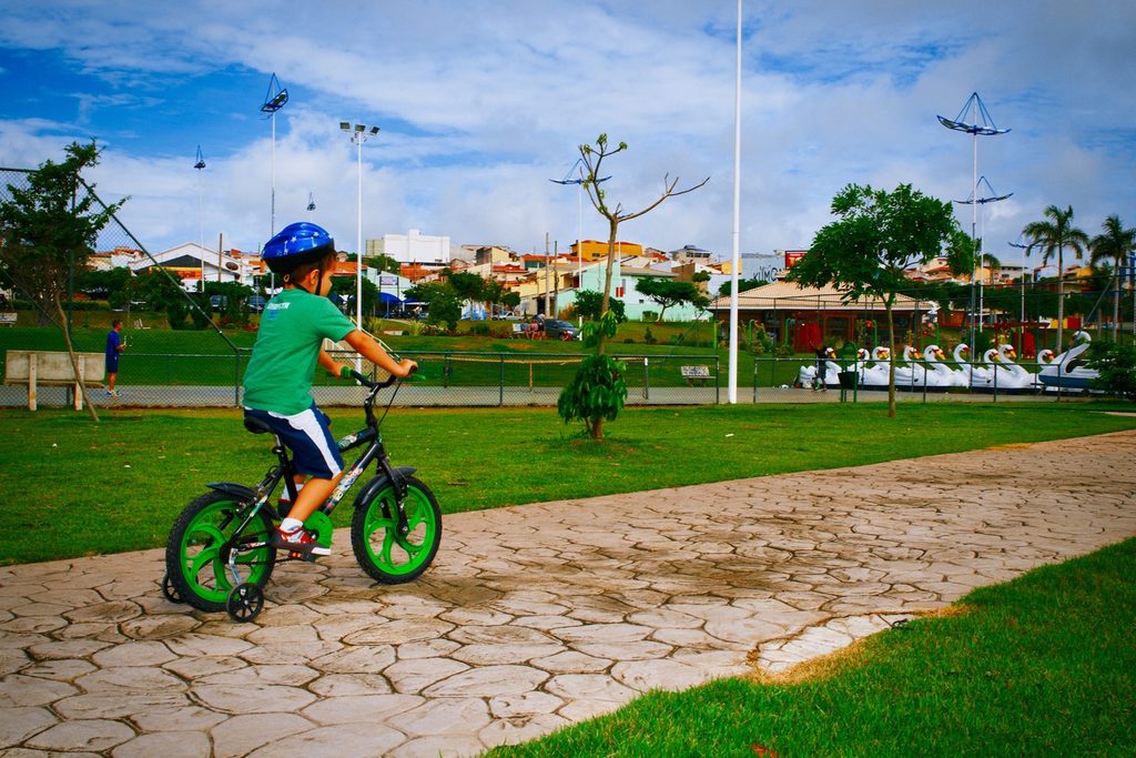Menino de capacete andando de bicicleta numa calçada do Parque Ecológico de Indaiatuba, com pedalinhos de cisne no lago ao fundo