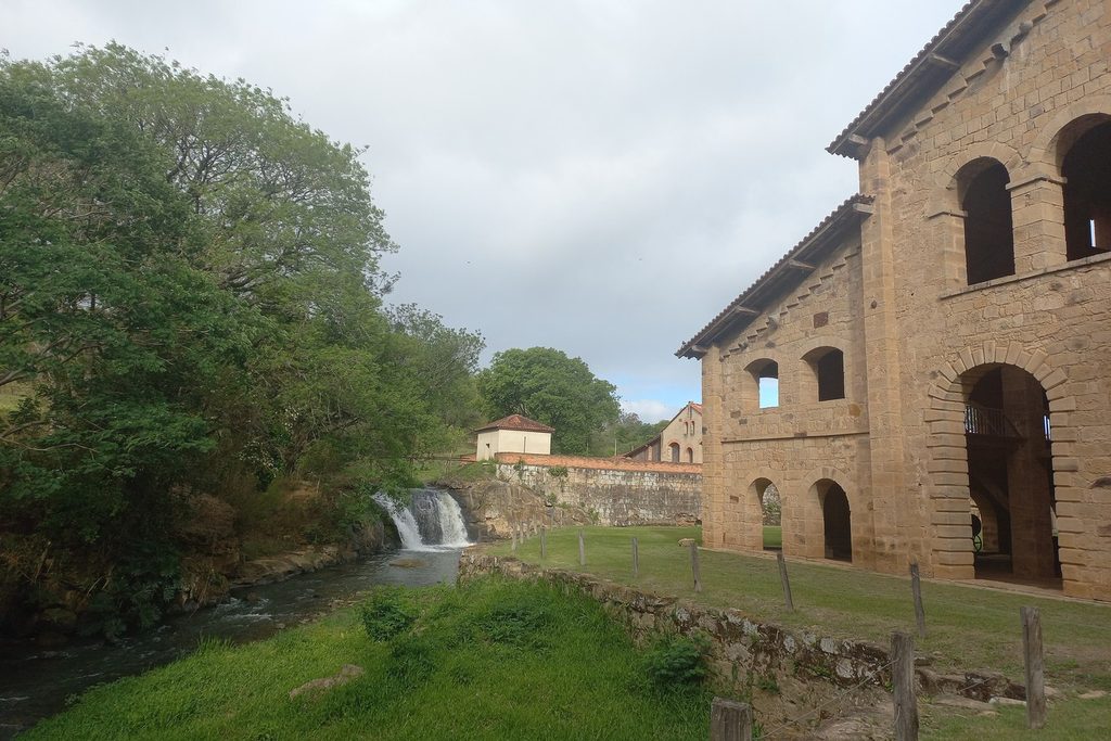 Prédio de pedra com janelas em arco ao lado de uma pequena cachoeira que desce da barragem entre a mata