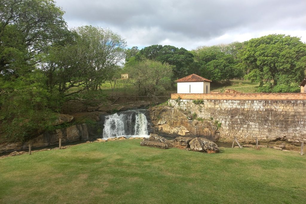 Pequena cachoeira descendo pelo muro de pedra da barragem, com gramado na frente e casinha branca no alto