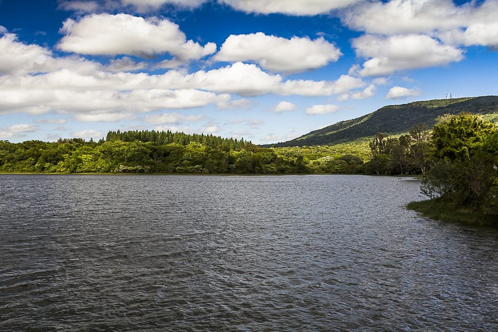 Represa de águas escuras com margens de mata e o Morro Araçoiaba ao fundo, sob céu azul com nuvens, na Fazenda Ipanema