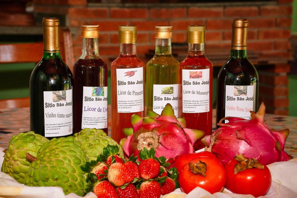 Garrafas de vinho e licores artesanais do Sítio São José, em Itatiba, com morangos, caquis, pitaias e pinhas na frente