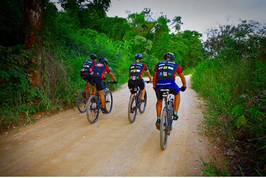 Grupo de ciclistas pedalando numa estrada de terra cercada de mata, no circuito de cicloturismo
