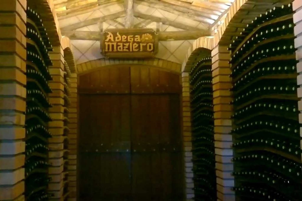Corredor de adega com arcos de tijolo e garrafas de vinho deitadas nas paredes, com a placa Adega Maziero sobre a porta de madeira