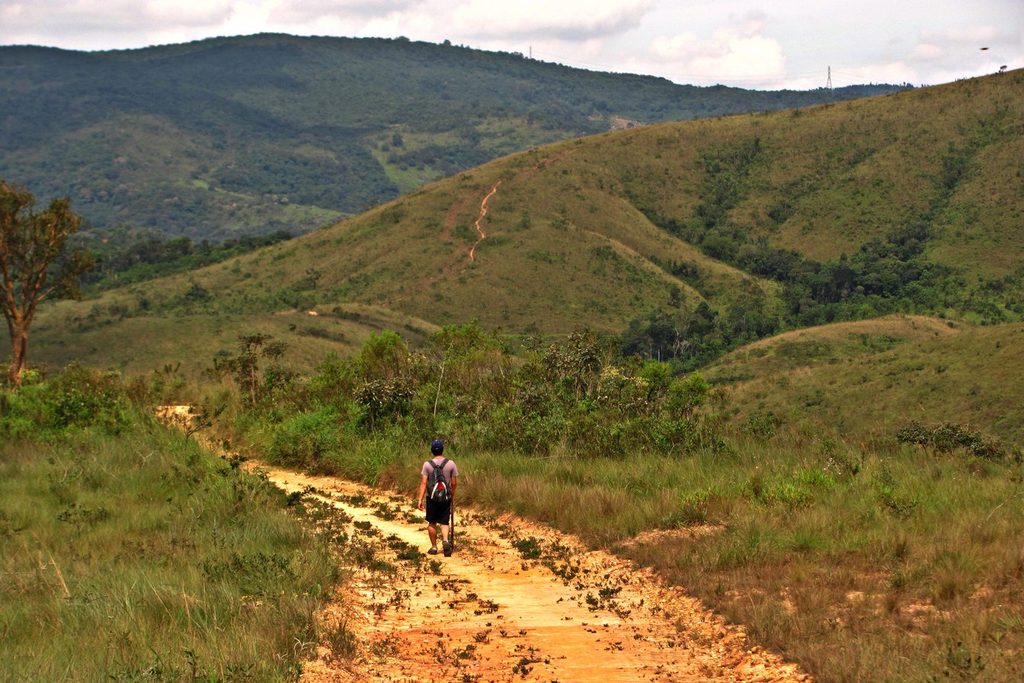 Visitante caminhando numa trilha de terra do Parque do Juquery, entre morros de capim e árvores baixas do cerrado