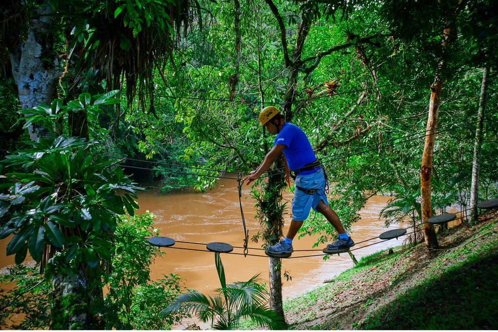 Homem de capacete atravessando uma ponte de arvorismo entre as árvores, sobre o Rio Juquiá, em Juquitiba