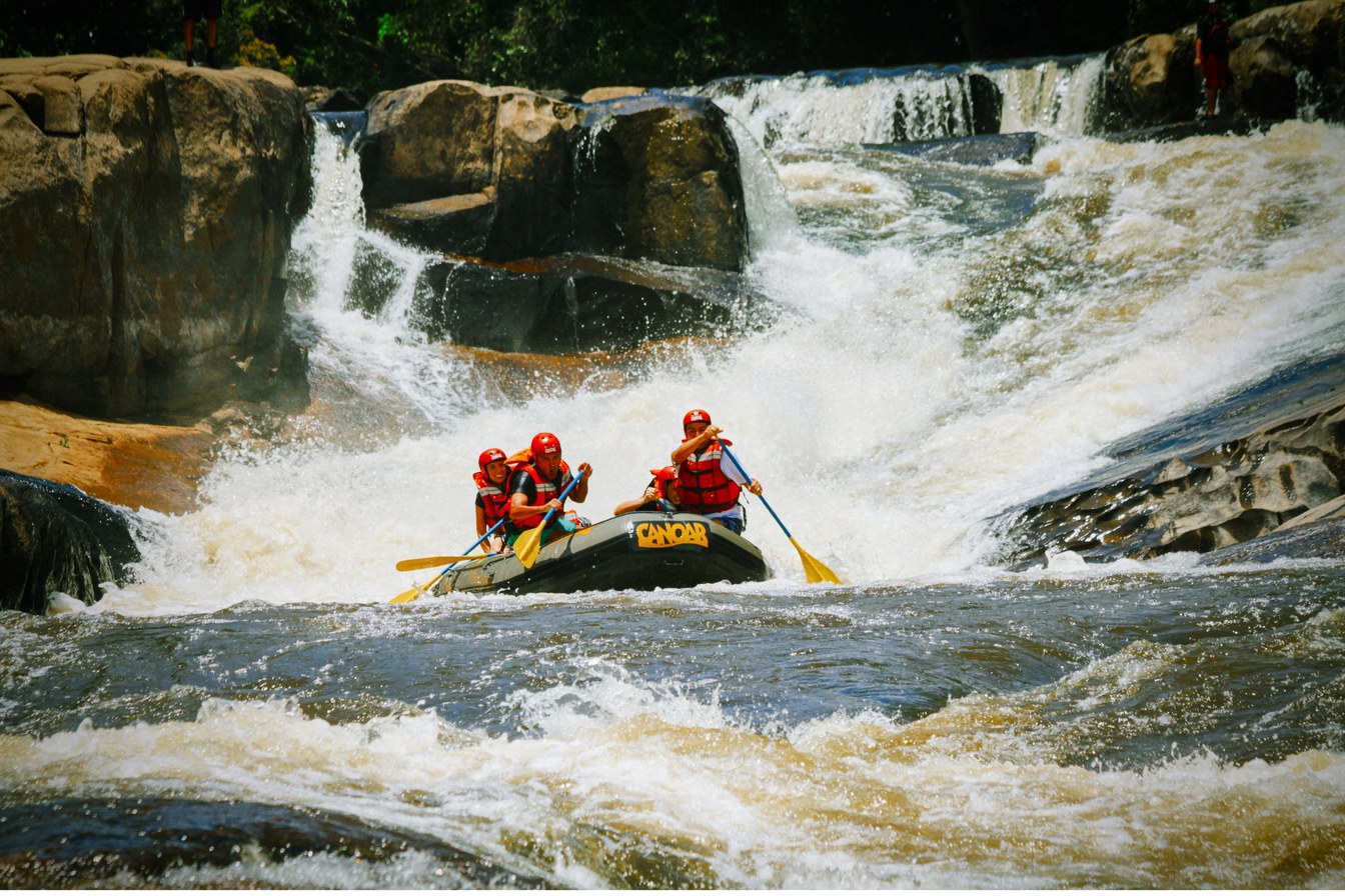 Bote de rafting com quatro pessoas de capacete vermelho descendo uma corredeira entre pedras em Juquitiba