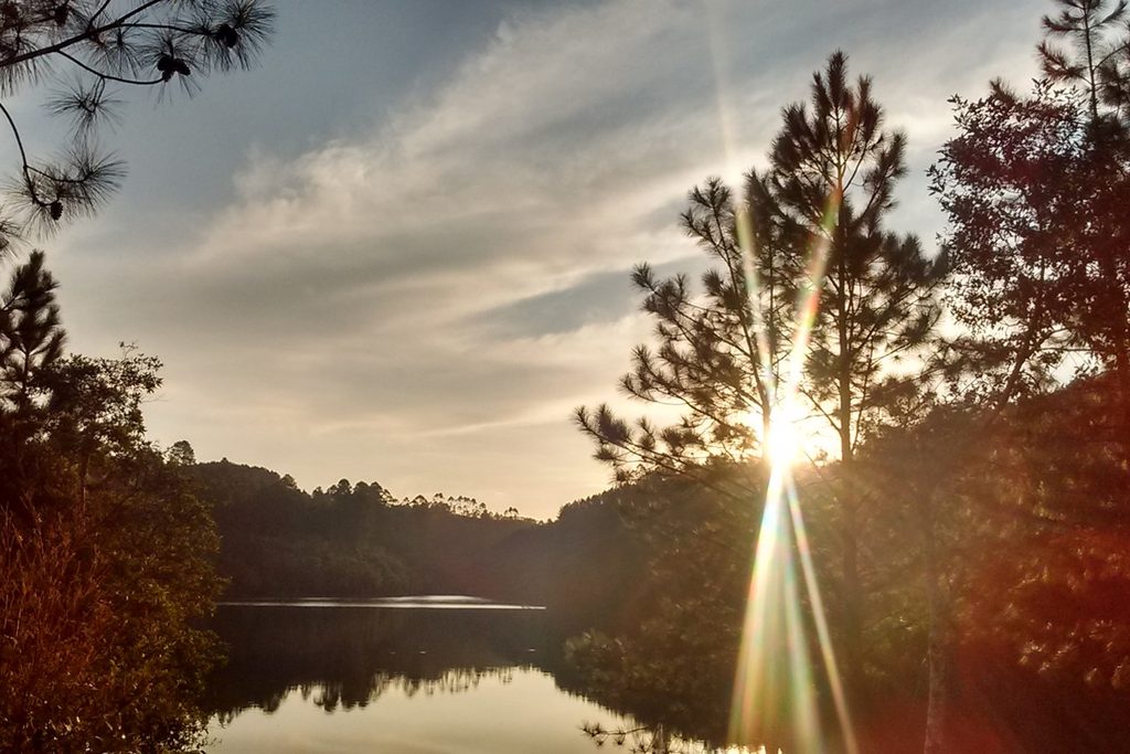 Pôr do sol sobre uma represa cercada de pinheiros e mata no bairro Meia Légua