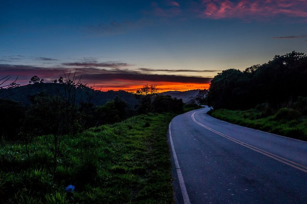 Estrada asfaltada em curva entre morros e mata, com o céu vermelho do pôr do sol