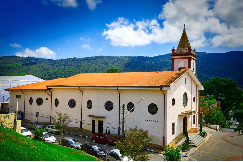 Igreja Matriz de Nossa Senhora do Desterro, em Mairiporã, branca com janelas redondas e torre, com a serra ao fundo