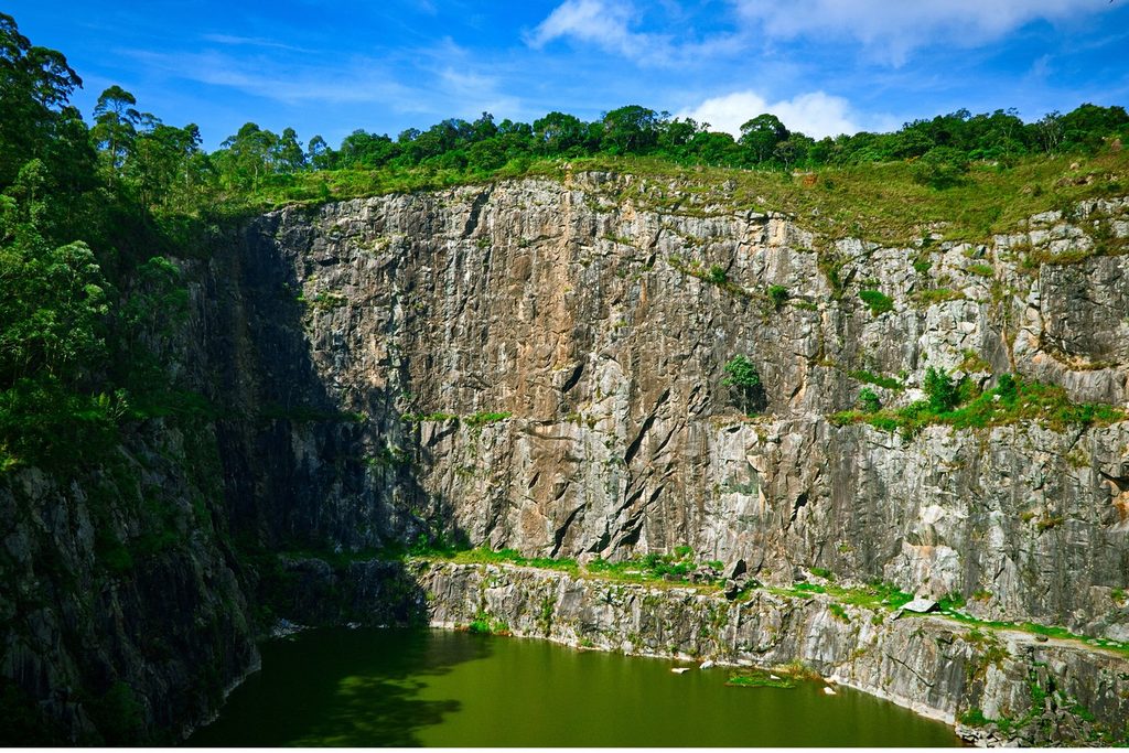 Paredão de pedra cinza de uma antiga pedreira em Mairiporã descendo até um lago verde, com mata no alto