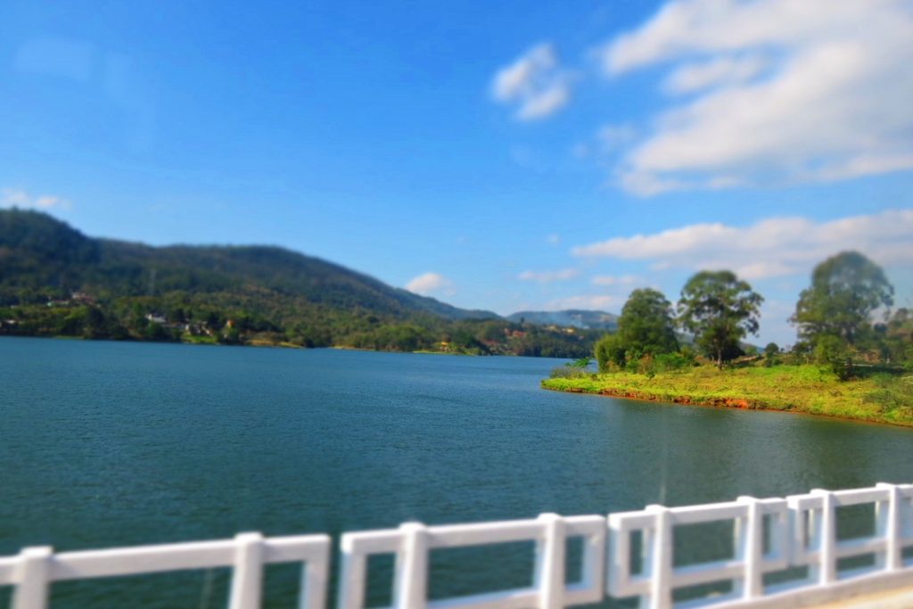 Represa de águas azuis em Mairiporã cercada de morros verdes, vista da mureta branca de uma ponte