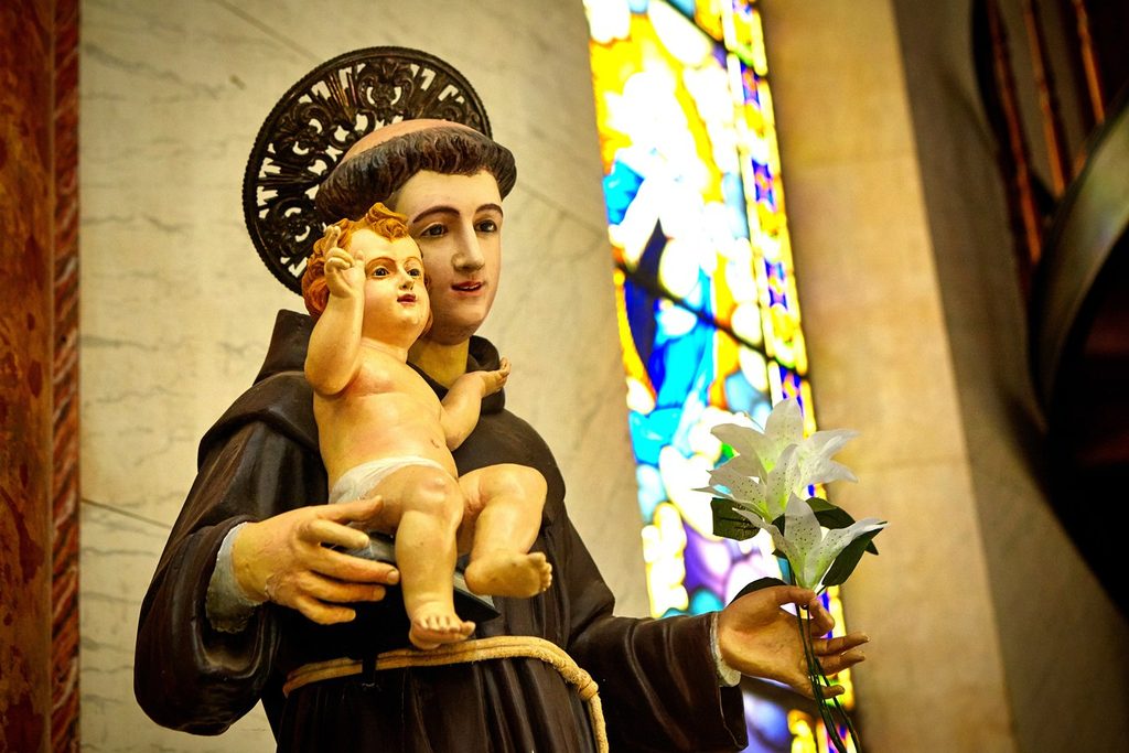 Imagem de Santo Antônio com o Menino Jesus no colo e lírios na mão, ao lado de um vitral colorido