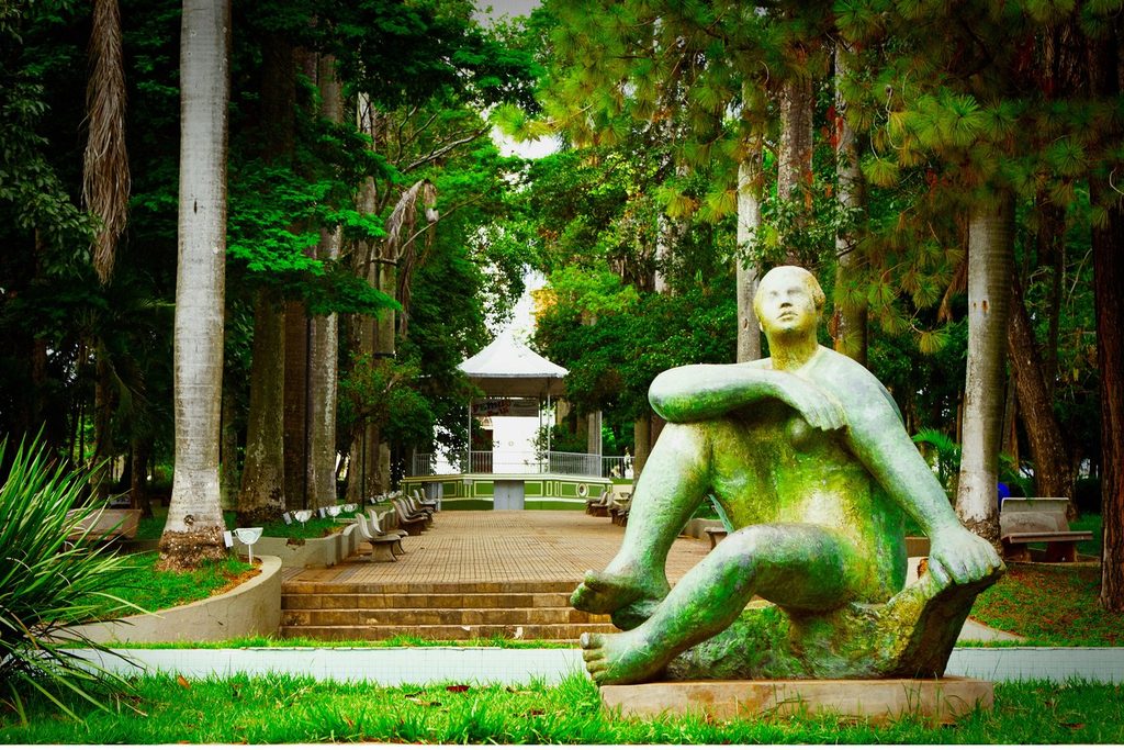 Escultura de bronze de mulher sentada com os braços sobre os joelhos na Praça Marechal Deodoro, em Mococa, com palmeiras e coreto ao fundo